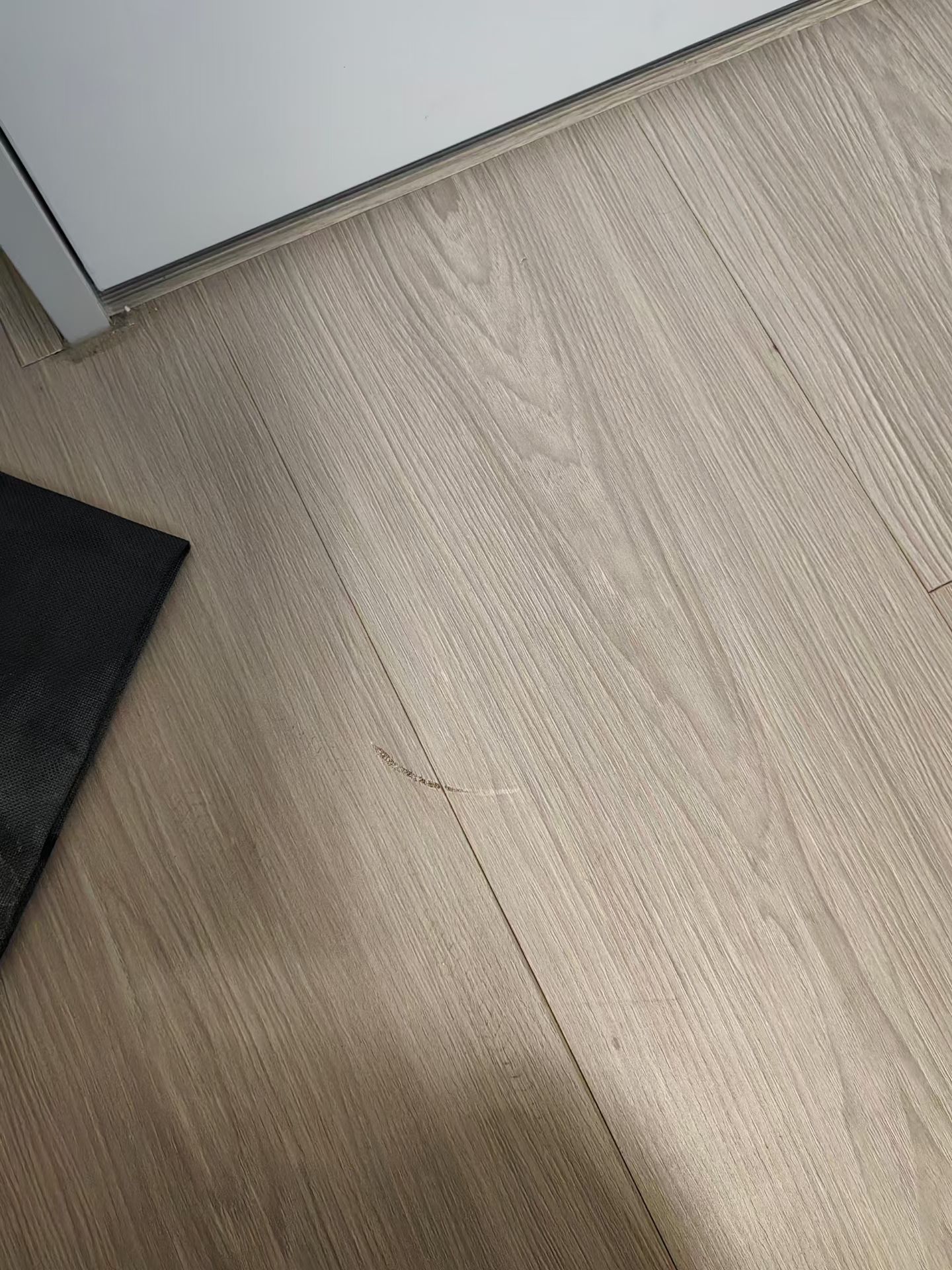 Wood-Grain Floor Scratch Repair — Before: A visible scratch across the wood-grain flooring near the doorway.
